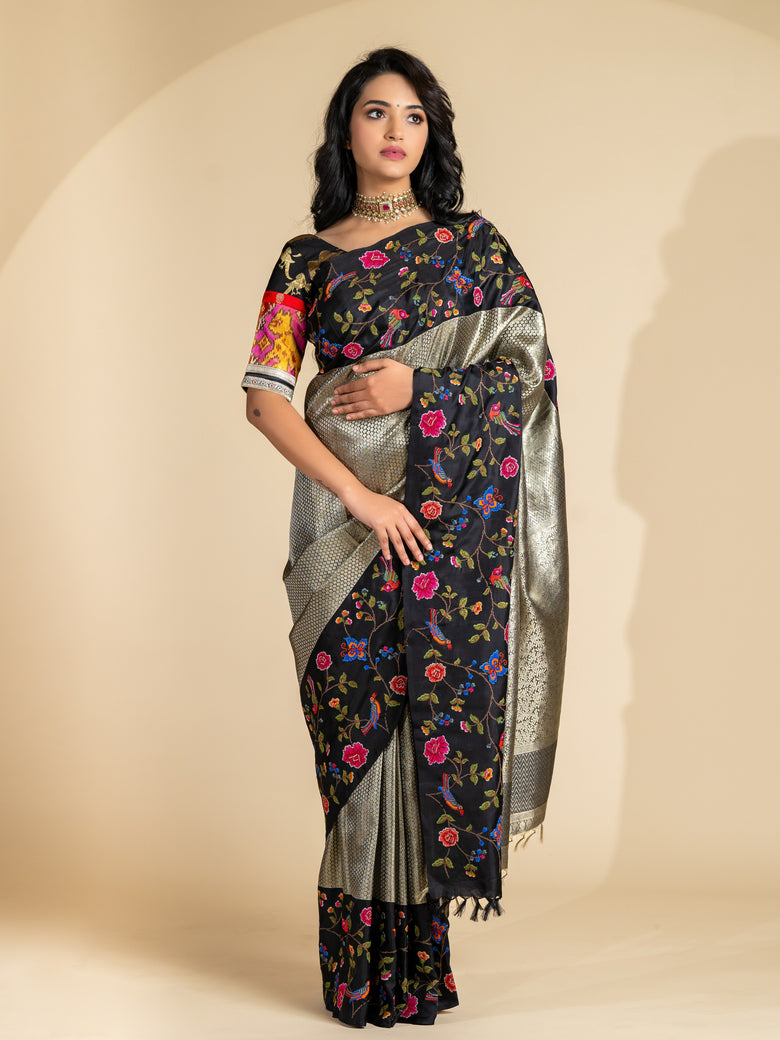 A woman modeling a black tissue kanjeevaram silk saree with a silver zari brocade design and floral and bird motifs on the border, along with a matching blouse piece.